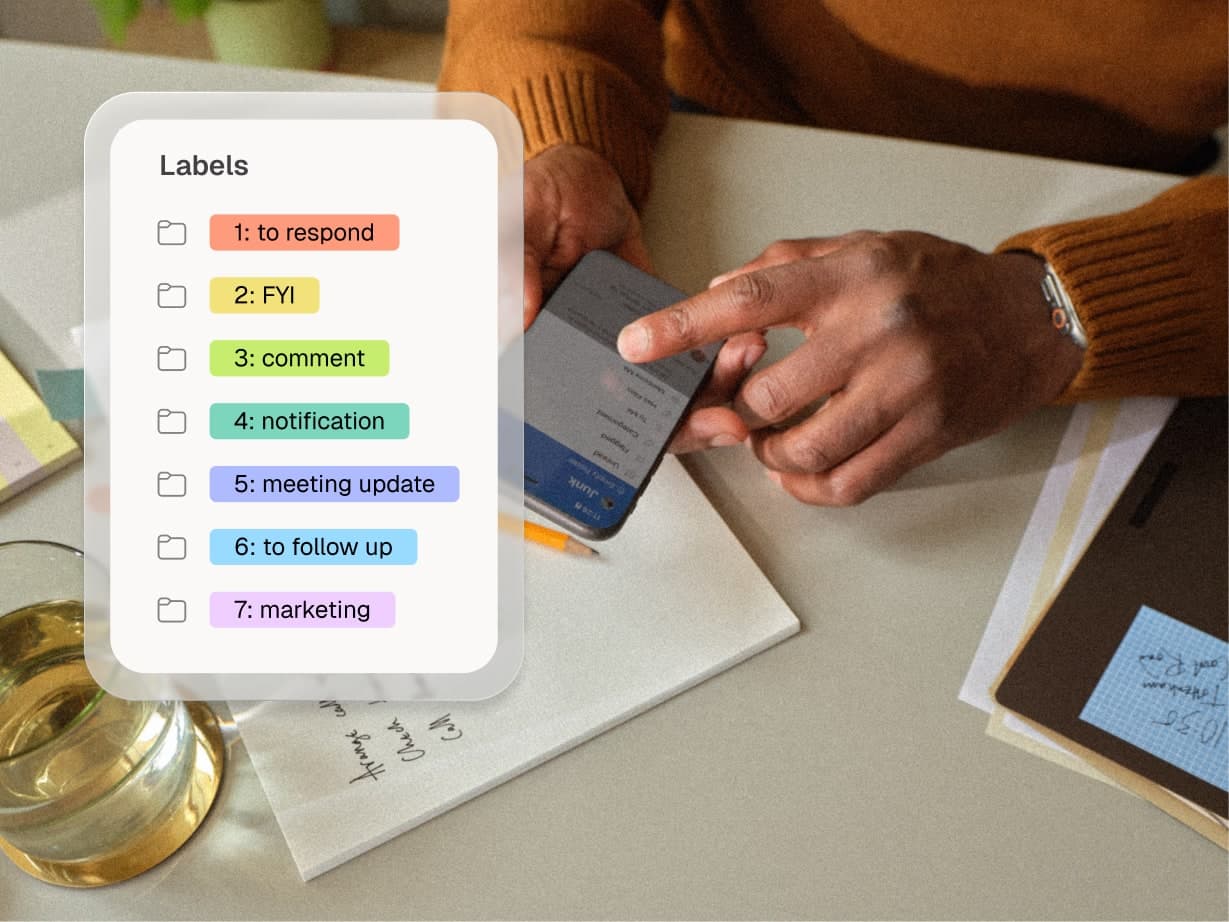 A person uses a smartphone at a table with labeled folders for tasks, a glass of water, papers, and a notebook with sticky notes.